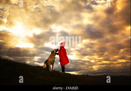 Kind im roten cape, das Deutschen Schäferhund unter sonnigem Himmel streichelt Stockfoto