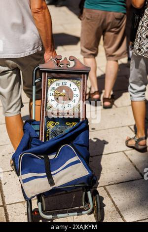 Ein Mann läuft durch einen Marktplatz und zieht einen Einkaufswagen mit einer alten hölzernen Uhr, die rausrastet. Stockfoto