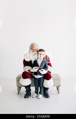 Weihnachtsmann sitzt auf der Bank mit einem Jungen im Anzug, minimalistischer weißer Rücken Stockfoto