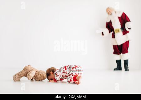 Baby im Weihnachts-Pyjama mit Teddybär, Weihnachtsmann im Hintergrund, Stockfoto