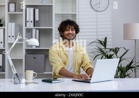 Ein lächelnder junger Mann an seinem Schreibtisch mit einem Laptop in einer hellen und modernen Büroumgebung. Konzept von Arbeit, Glück und Produktivität. Stockfoto