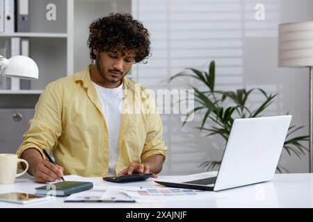 Junger Mann, der im Heimbüro mit Rechner und Laptop arbeitet, Dokumente analysiert und Notizen macht. Stockfoto