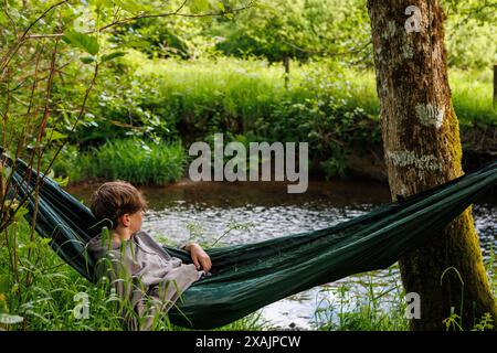 Ein junger Teenager entspannt sich, während er aus einer Hängematte blickt, die zwischen zwei Bäumen am Ufer eines kleinen Flusses hängt. Stockfoto