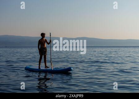 Ein junger, sportlich aussehender Teenager steht auf einem Paddelbrett im goldenen Sonnenlicht mit Bergen im Hintergrund, Omis Croatia. Stockfoto