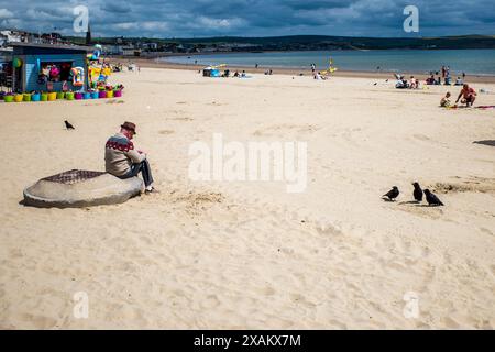 Einsamer Mann an einem fast leeren Strand mit drei Krähen, die ihn anschauen. Weymouth Beach, Dorset. Einsamkeit, Denken, Kommunizieren. Vögel. Stockfoto