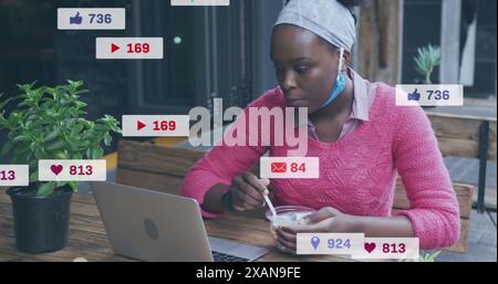 Bild einer Social-Media-Ikone über eine afroamerikanische Frau, die einen Snack trinkt und in einem Café einen Laptop benutzt Stockfoto