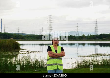 Porträt einer Umweltingenieurin, die stolz gegen ein Kraftwerk, Hochspannungsmast und Reservoir im Hintergrund lächelt. Energie, Ökologie Stockfoto