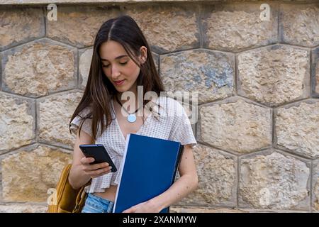 Ein junges College-Mädchen lehnt sich an eine Wand, Rucksack und Bücher in der Hand, überprüft ihr Smartphone. Moderner Student, akademisches Leben, Technologie und College Stockfoto