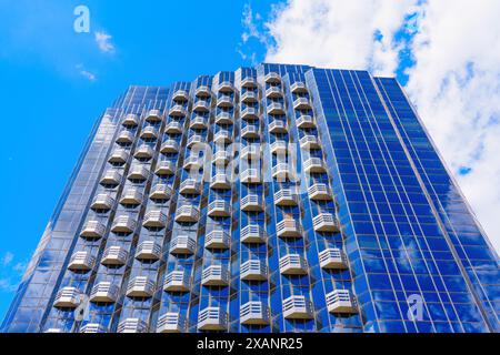 Beeindruckender Blick nach oben auf ein Hochhaus mit einer Glasfassade und Reihen identischer Balkone, alle vor einem hellblauen Himmel. Stockfoto