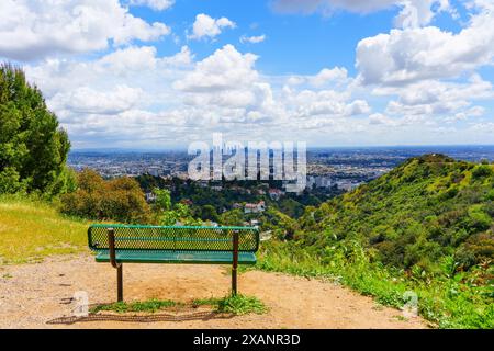 Panoramablick vom Runyon Canyon, mit einer einsamen grünen Bank auf einem unbefestigten Weg mit Blick auf die weitläufige Landschaft mit üppigem Grün und die Dista Stockfoto