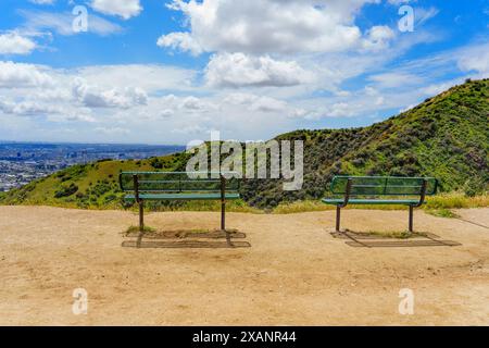 Zwei grüne Bänke auf dem Runyon Canyon Trail mit Blick auf die weitläufige Stadtlandschaft von Los Angeles. Die Bänke stehen vor dem Hintergrund des üppigen gre Stockfoto