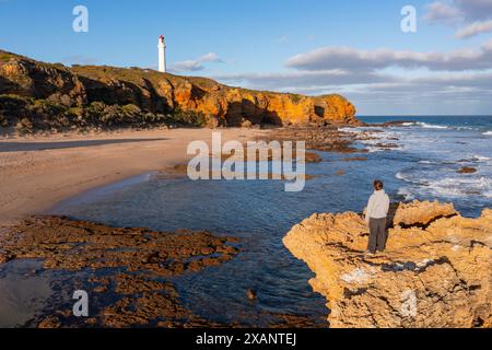 Ein Mann auf einem Felsvorsprung, der auf einen Leuchtturm auf einer Klippe in der Ferne am Aireys Inlet an der Great Ocean Road in Victoria, Australien, blickt Stockfoto