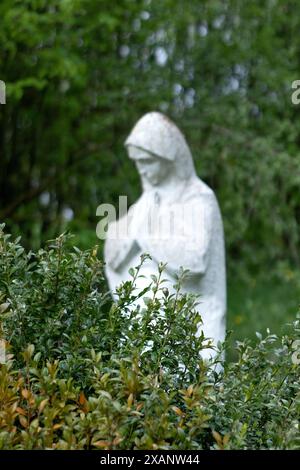 Eine beschädigte, alte Statue der Mutter Gottes, verlassen in der Friedhofskippe, verwischte Bäume mit grünen Blättern im Hintergrund. Stockfoto