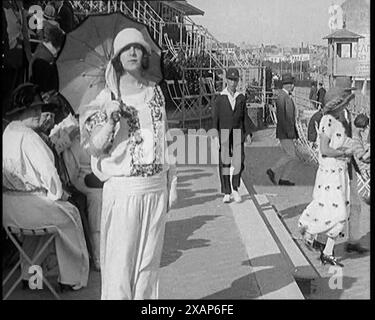 Weibliche Zivilist mit intelligentem Outfit und Hut mit einem Sonnenschirm, der während eines Pferderennens in Richtung Kamera geht, 1920er Aus „Time to Remember – Teenage Flapper“, 1920er Jahre (Reel 2); ein Dkumentary über das Leben von Frauen in den 1920er Jahren – großartiger Kommentar von Joyce Grenfell. Stockfoto