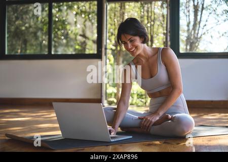 Glückliche Yogatrainerin der jungen Frau, die im Yoga-Studio sitzt und einen Laptop benutzt. Stockfoto