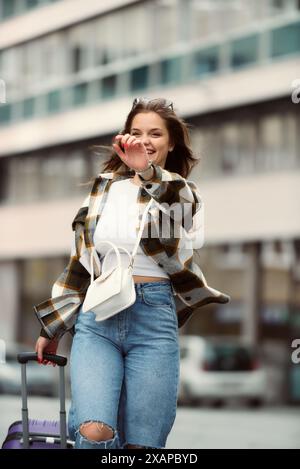 Charmante Frau schlendert in einem stilvollen karierten Hemd, einer blauen Hose und einer Sonnenbrille durch die Stadt. Stockfoto