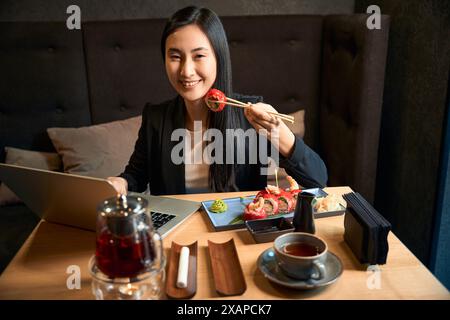 Positive asiatische Frau, die Sushi mit Essstäbchen hält und mit Laptop auf dem Tisch sitzt Stockfoto