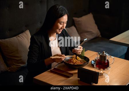 Positive attraktive Frau beim Mittagessen und beim Chatten auf dem Smartphone Stockfoto