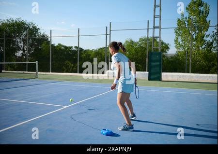 Eine Frau in den 40ern übt Tennis auf einem Outdoor Court. Sie trägt Tenniskleidung und konzentriert sich auf den Ball. Der Hintergrund zeigt ein c Stockfoto