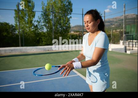 Eine Frau in den 40ern übt ihre Tennisfähigkeiten auf einem Outdoor-Platz aus. Sie konzentriert sich auf Ball und Schläger, trägt Sportkleidung und Armbänder o Stockfoto