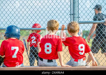 Eine Reihe junger T-Ball-Jungs, die auf der Bank hinter dem Heimteller sitzen und darauf warten, zu schlagen. Stockfoto