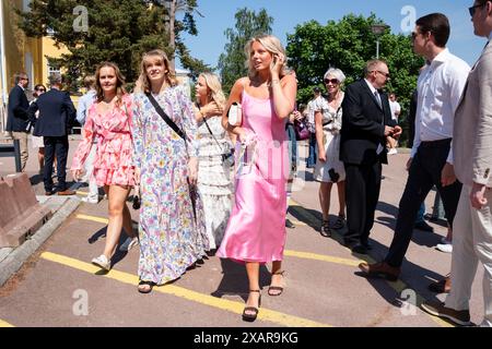 FREUNDE IN SOMMERKLEIDERN WARTEN AUF DIE ABSCHLUSSFEIER, MARIEHAMN: Familien und Freunde treffen sich vor dem Hauptschulgebäude zum Åland-Abschlussfeiertag 2024 im Åland Lyceum (Ålands Gymnasium) in Mariehamn, Åland Archipel, Ostsee, Finnland – an diesem Tag verlassen die Schüler die Schule zum letzten Mal. Im Rahmen dieser nordischen Tradition werden Plakate mit Babybildern der Absolventen aufgeklebt und die Studenten selbst tragen ihre Studentmössa im Matrosenstil und kleiden sich in Anzügen (Jungen) und weißen Kleidern (Mädchen). Foto: Rob Watkins. Stockfoto