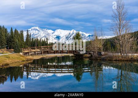 Holzbogen Fußgängerbrücke in Cascade Teiche Park im Herbst sonnigen Tag, schneebedeckten Mount Astley Reflexion auf der Wasseroberfläche. Banff National Park Stockfoto