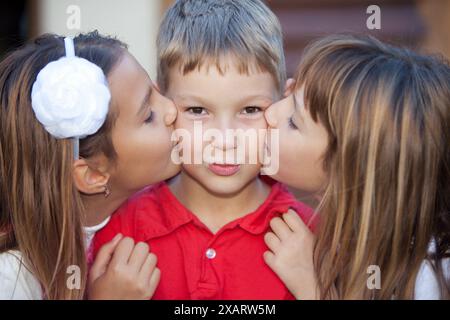 Porträt von drei glücklichen Kindern. Zwei Mädchen küssen einen überraschten Jungen auf die Wangen Stockfoto