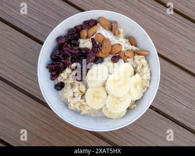 Ein Blick von oben auf eine weiße Schüssel Haferbrei mit frischen Bananenscheiben, knusprigen Mandeln und süßen getrockneten Preiselbeeren auf einem rustikalen Tisch Stockfoto