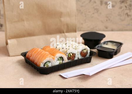 Plastikteller mit leckeren Sushi-Rollen, Essstäbchen und Papiertüte auf beigem Grunge-Hintergrund. Lieferkonzept Stockfoto