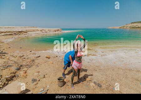 Totes Meer, Jordanien - 19. April 2024: Die Frau steht am Ufer des Toten Meeres, als ein Mann sie mit Schlamm bedeckt. Beide genießen den heißen Sommer Stockfoto