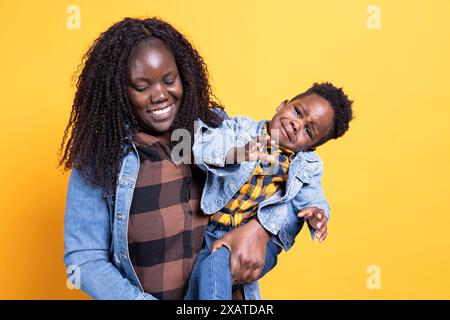 Ängstlich aufgeregt, süßes Kind fängt an zu weinen vor gelbem Hintergrund, seine Mutter trägt ihn und bringt ihn zum Lächeln. Die afroamerikanische Mutter versucht, ihr Kleinkind zu beruhigen. Stockfoto