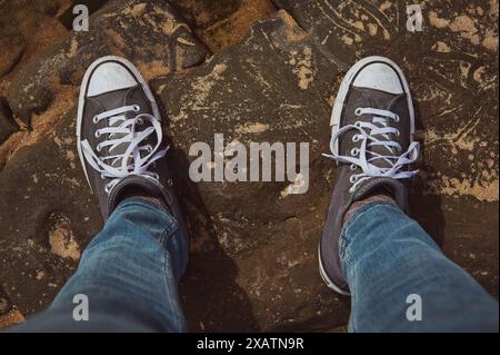 Nahaufnahme einer Person mit lässigen Sneakers und blauen Jeans, die auf einem felsigen Untergrund steht. Das Bild zeigt die Einfachheit und Robustheit eines Outdos Stockfoto