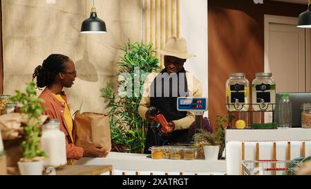 Regelmäßiger Kunde, der sich mit Bio-Artikeln zur Kasse nähert, bereitet sich auf den Kauf von Naturprodukten und hausgemachten Saucen aus dem Bio-Supermarkt vor. Junge Frau fördert nachhaltigen Lebensstil. Kamera 1. Stockfoto