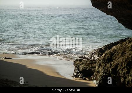 Eine ruhige Strandszene mit sanften Wellen, die gegen die Sandküste plätschern, eingerahmt von großen Felsformationen auf der rechten Seite. Stockfoto