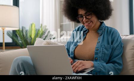 Afroamerikanische Frau ethnische Mädchen mit Laptop zu Hause chatten mit Freunden in sozialen Medien SMS mit Computer erstaunte Nachrichten lächelnd glücklich Stockfoto