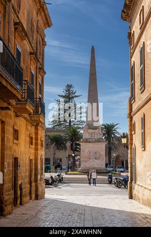 Ciutadella, Spanien - 26. Januar 2024: Enge Kopfsteinpflasterstraße im historischen Zentrum von Ciutadella mit dem Obelisken Ciutadella im Hintergrund Stockfoto