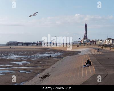 Menschen sitzen auf der Promenade neben dem Sandstrand und Blackpool Tower in der Stadt Blackpool, Lancashire, England. Juni 2024 Stockfoto