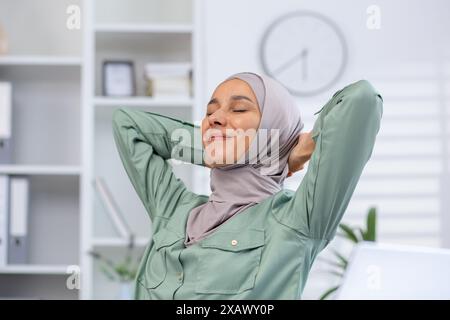 Frau im Hijab dehnt sich und entspannt am Arbeitsplatz im Büro mit einem glückseligen Ausdruck. Konzept der Work-Life-Balance, Wohlbefinden und Entspannung Stockfoto