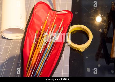 Verschiedene Pinsel in einem roten Etui und Abdeckband auf einem Tisch, bereit für einen Kunstkurs. Perfekt für Kreativität und Workshop-Themen. Stockfoto