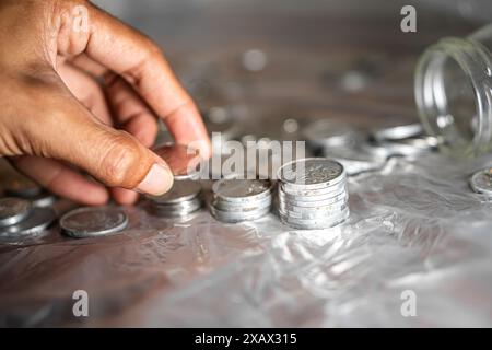 Eine Nahaufnahme einer Hand, die Silbermünzen auf einer transparenten Oberfläche stapelt, mit weiteren verstreuten Münzen. Ein Glasgefäß ist teilweise im Hintergrund sichtbar Stockfoto