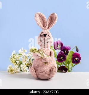 Gestrickter rosafarbener Hase mit weißem Flieder und Blumenstrauß der Frühlingsblumen von Flieder und Bratsche auf blauem Hintergrund. Das Konzept des Frühlings, Ostern, Muttertags Stockfoto