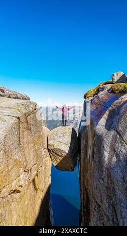 Eine Person steht mit ausgestreckten Armen auf einer Felsformation in Kjeragbolten, Norwegen, an einem klaren Tag. Asiatische Frauen mit erhobenen Händen in Kjeragbolten, Norwegen Stockfoto