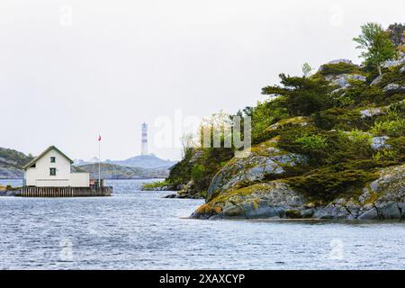 Weiße Strandhütte auf Flekkeroya mit Leuchtturm von Oksoy im Hintergrund. Reisetrend für kalte Reiseziele. Kristiansand, Norwegen Stockfoto