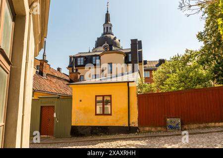 Der Turm und die Kuppel der Katarina Kyrka erheben sich über einem kleinen gelben Holzhaus in einer engen Gasse im Södermalm-Bezirk. Der Architekt Jean de la Vallée entwarf die barocke Kirche, deren Bau 1656 begann und 1695 abgeschlossen wurde. Ein Brand zerstörte die Kirche 1723 fast vollständig, woraufhin sie mit einem größeren, achteckigen Turm wiederaufgebaut wurde. Ein zweiter Großbrand im Jahr 1990 ließ kaum mehr als die Außenwände stehen, und seit ihrer Wiedereröffnung im Jahr 1995 dient die Kirche wieder als Pfarrkirche. Södermalm, Stockholm, Stockholms län, Schweden. Stockfoto