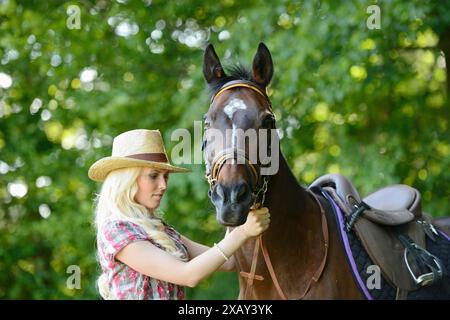 Frau (Cowgirl) in Strohhut und kariertem Hemd steht neben einem polnisch-arabischen Pferd auf einem Feld in Bayern Stockfoto