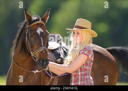 Frau (Cowgirl) in Strohhut und kariertem Hemd steht neben einem polnisch-arabischen Pferd auf einem Feld in Bayern Stockfoto