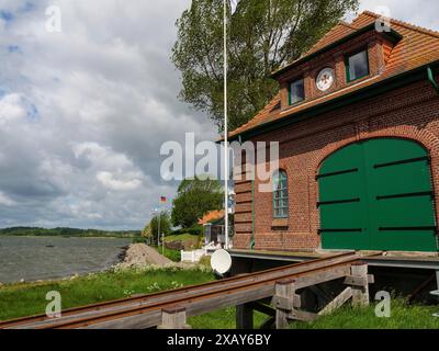 Rotes Backsteingebäude mit grünem Tor an der Uferpromenade mit Bahngleisen und Fahnenmasten, Maasholm, Schleswig-Holstein, Deutschland Stockfoto