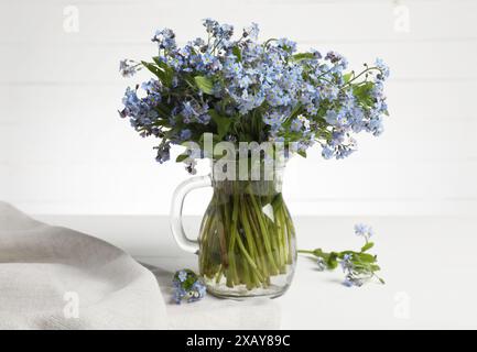 Blumenstrauß mit wunderschönen Vergissmeinsblumen in Glaskrug und blauem Tuch auf weißem Tisch Stockfoto