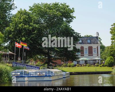 Ein Touristenboot auf dem Kanal, umgeben von grünen Bäumen und eine historische Villa mit Fahnen im Vordergrund, friedrichstadt, schleswig-holstein, deutschland Stockfoto